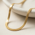 Close up of a 18k gold plated thick, flat herringbone chain necklace resting on a textured white surface.