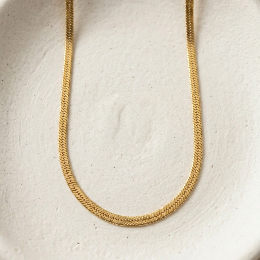 18k gold plated thick, flat herringbone chain necklace resting on a textured white surface.