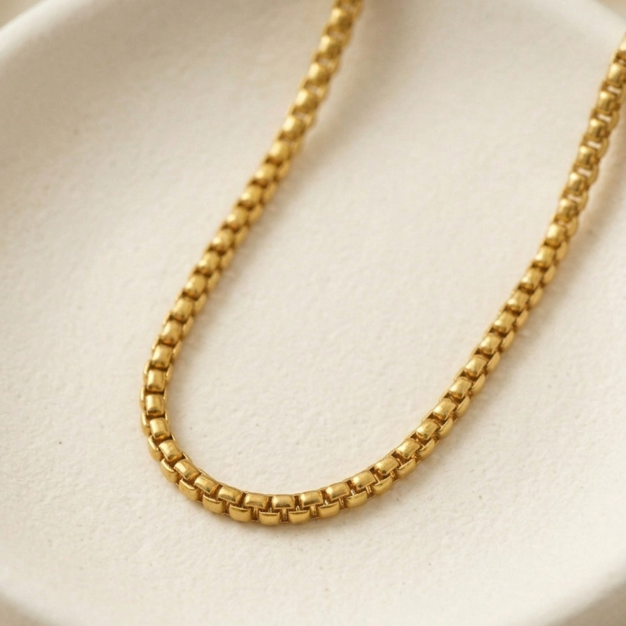 A close-up of a gold box chain necklace draped over a round, textured cream-colored display stand.
