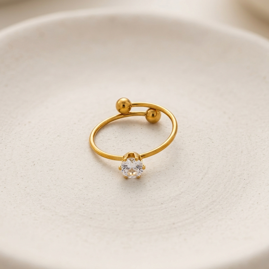 an 18k gold plated minimalist adjustable ring featuring a single CZ diamond and two small gold spheres, resting on a matte white ceramic plate.