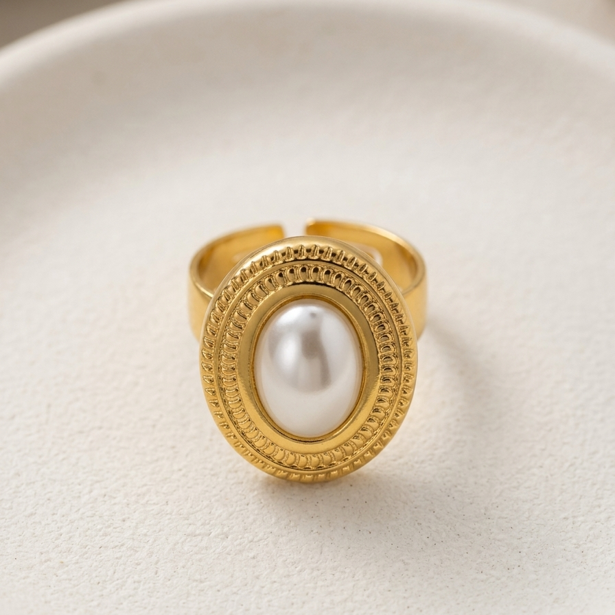 Close up of a premium 18k gold plated statement ring featuring a large oval pearl set in a detailed vintage bezel, displayed on a cream textured ceramic surface.