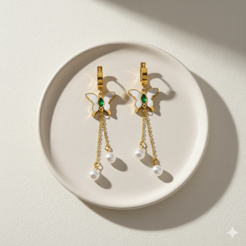 Gold butterfly huggie earrings with green stone center and dangling pearl chains on beige background.