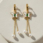Gold butterfly huggie earrings with green stone center and dangling pearl chains on beige background.