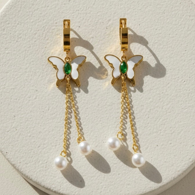 Gold butterfly huggie earrings with green stone center and dangling pearl chains on beige background.