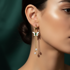 Gold butterfly huggie earrings with green stone center and dangling pearl chains on beige background.