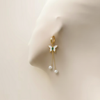 Gold butterfly huggie earrings with green stone center and dangling pearl chains on beige background.