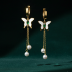 Gold butterfly huggie earrings with green stone center and dangling pearl chains on beige background.