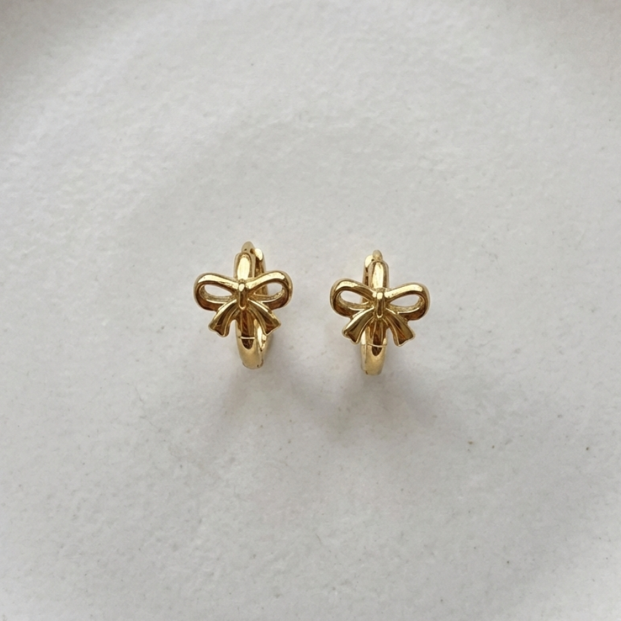 Gold ribbon bow huggie earrings on white ceramic plate.