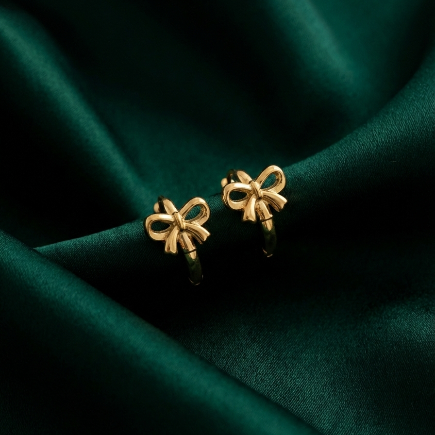 Gold ribbon bow huggie earrings on white ceramic plate.