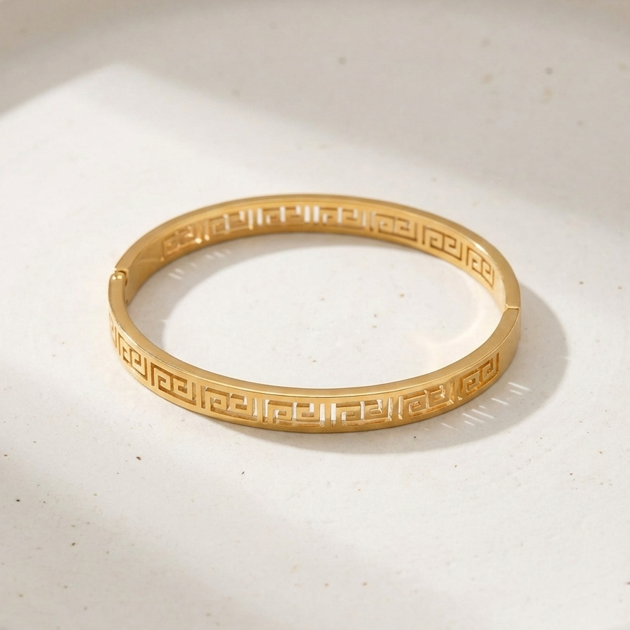 Gold oval bangle bracelet with cut-out Greek Key pattern on white plate.