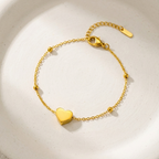 Dainty gold chain bracelet with small heart charm and beads on white stone background.