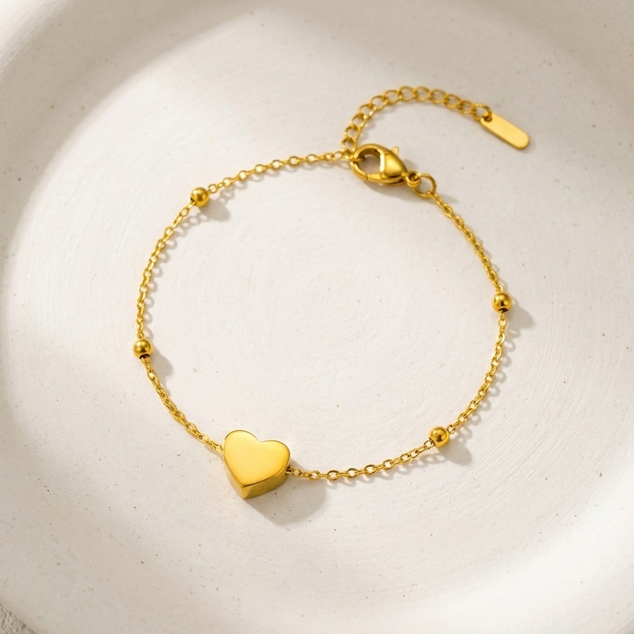 Dainty gold chain bracelet with small heart charm and beads on white stone background.