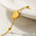 Dainty gold chain bracelet with small heart charm and beads on white stone background.