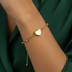 Dainty gold chain bracelet with small heart charm and beads on white stone background.