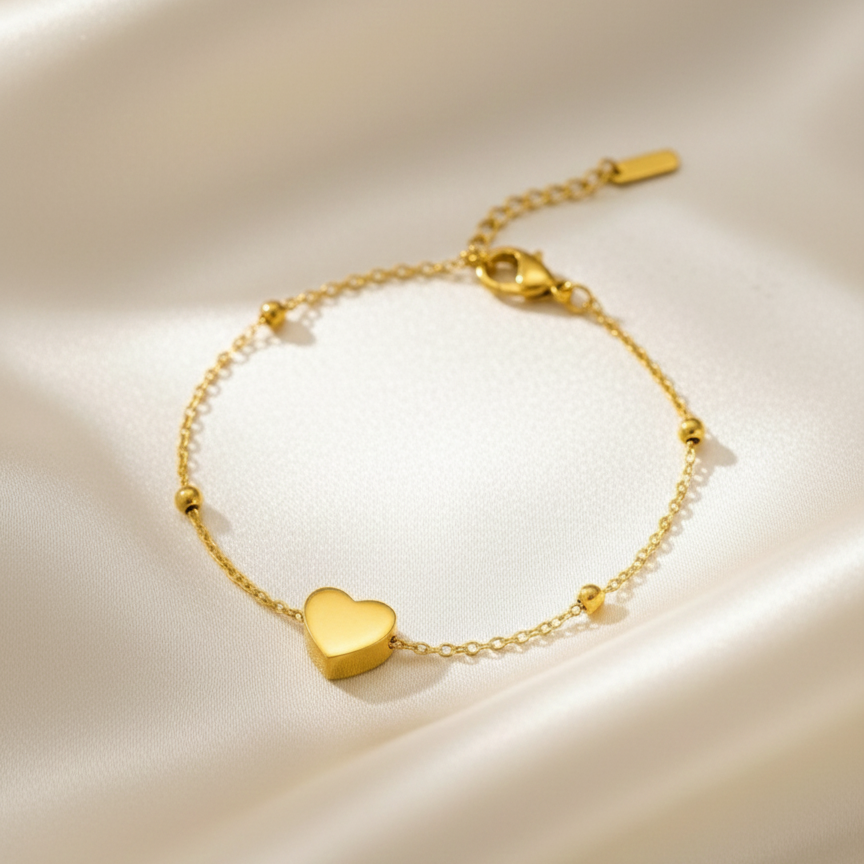 Dainty gold chain bracelet with small heart charm and beads on white stone background.