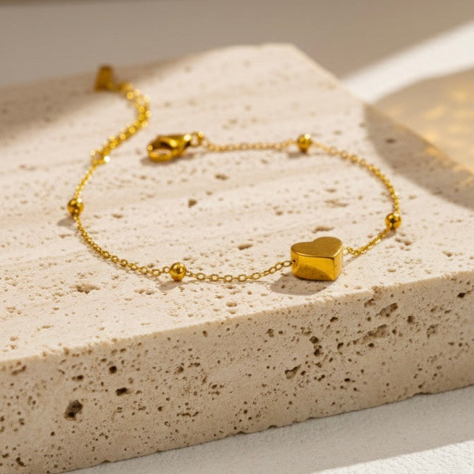 Dainty gold chain bracelet with small heart charm and beads on white stone background.