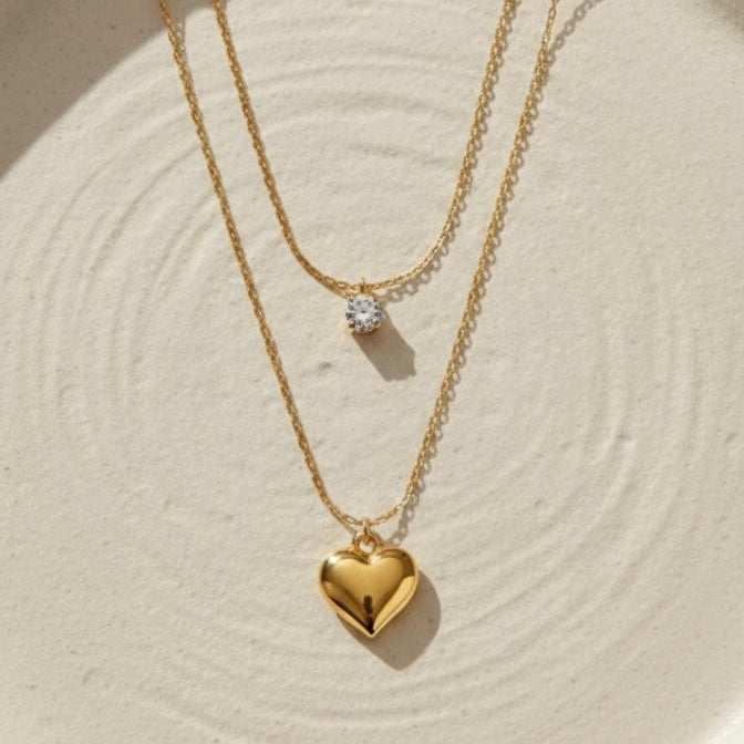Double layered gold necklace with crystal solitaire and puffy heart pendant on beige plate.