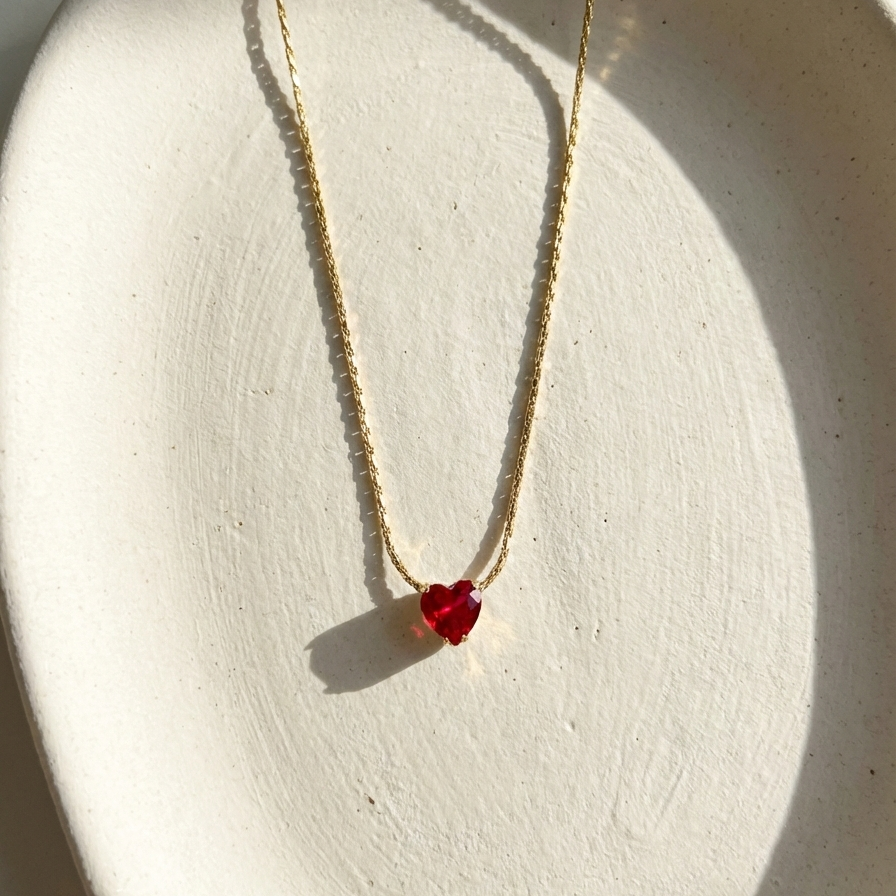 Gold necklace with faceted ruby red heart crystal pendant on white background.