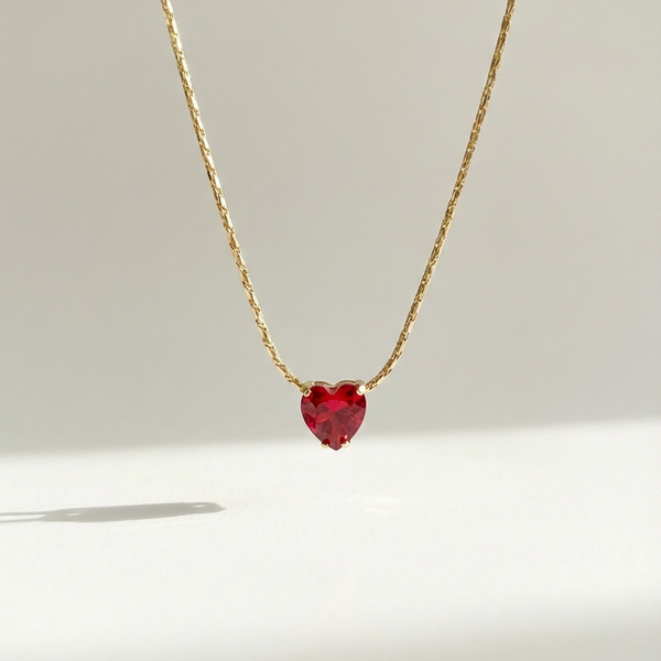 Gold necklace with faceted ruby red heart crystal pendant on white background.