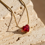 Gold necklace with faceted ruby red heart crystal pendant on white background.