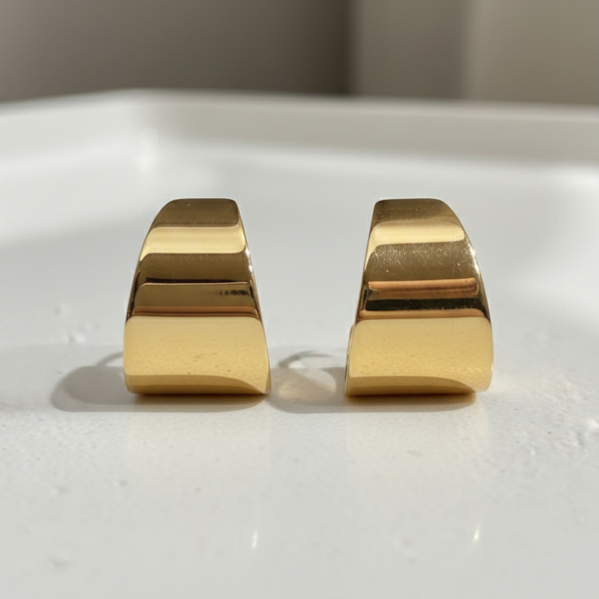 Wide sculpted gold huggie earrings on beige stone background.
