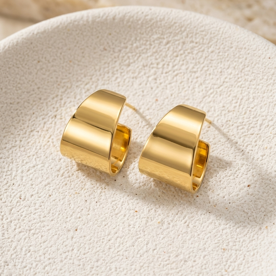 Wide sculpted 18kt gold plated huggies on a beige stone background.